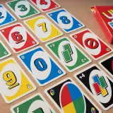 Karty do gry UNO, w tym karty z cyframi 0-9 i specjalne karty, leżą na drewnianym stole. Zestaw zawiera pudełko z grą. Zasady gry są proste: uno dos.