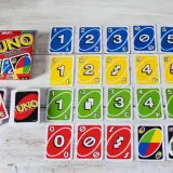 Karty do gry UNO ułożone w rzędy, w tym karty z numerami 0-5 w kolorach niebieskim, żółtym i zielonym, a także karty specjalne. Pudełko UNO i stos kart z numerem 9. Gra w UNO to świetna zabawa, ale czasem zdarza się uno blokada!