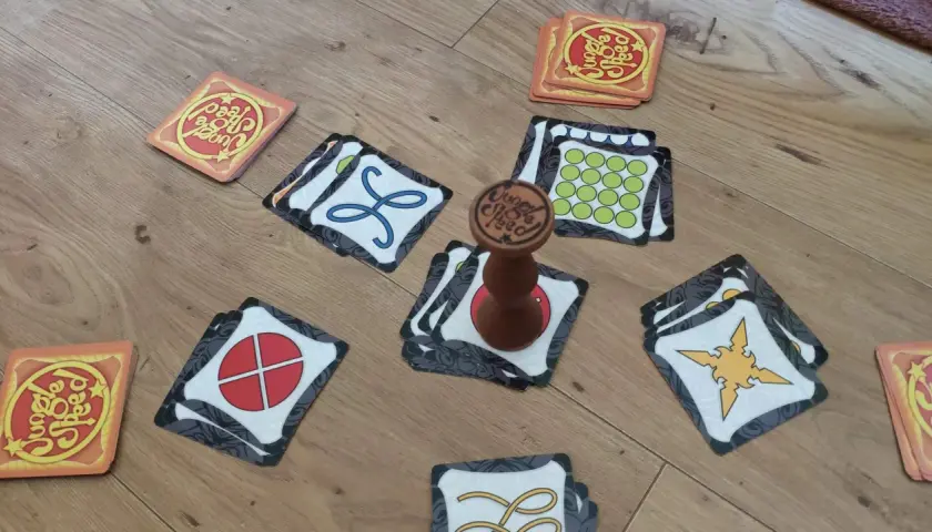 Karty do gry Jungle Speed leżą na drewnianej podłodze. Widać totem i różne symbole na kartach.