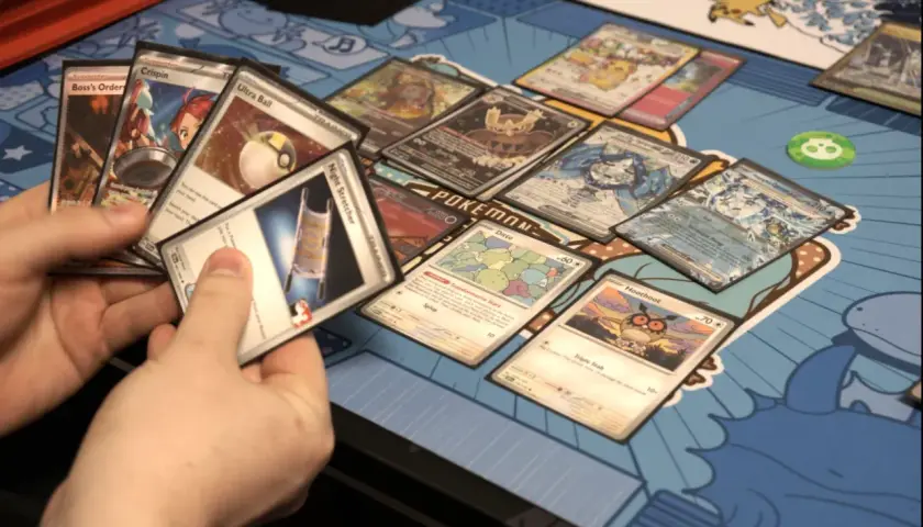 Ręce trzymają karty Pokémon, pokazując, jak grać w karty Pokémon. Widoczne karty to m.in. Boss's Orders, Ultra Ball i Ditto.