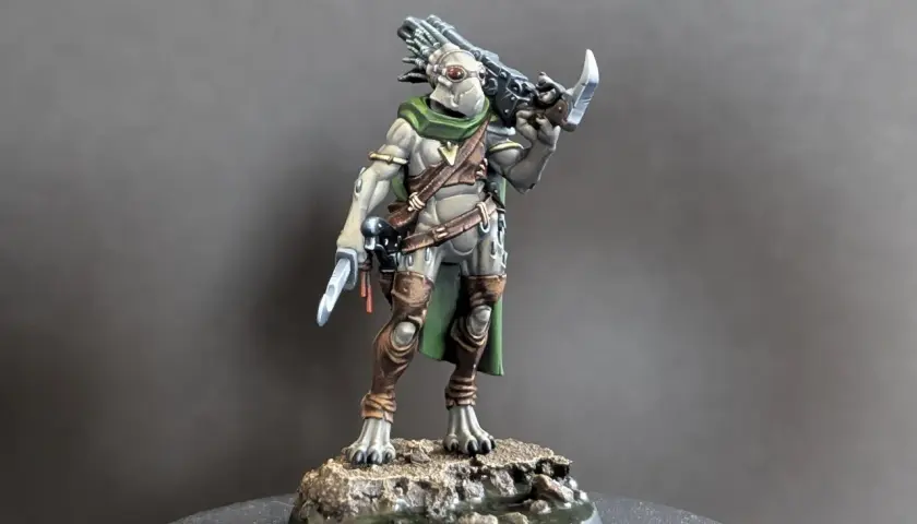 Zbrojny kroot w zielonej pelerynie i brązowych ochraniaczach, z nożem w dłoni i karabinem na plecach, gotowy do walki.