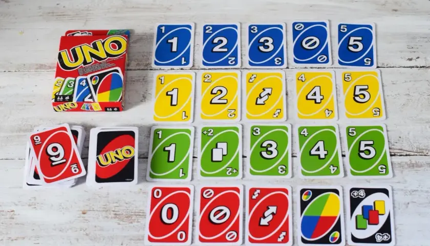 Karty do gry UNO ułożone w rzędy, w tym karty z numerami 0-5 w kolorach niebieskim, żółtym i zielonym, a także karty specjalne. Pudełko UNO i stos kart z numerem 9. Gra w UNO to świetna zabawa, ale czasem zdarza się uno blokada!